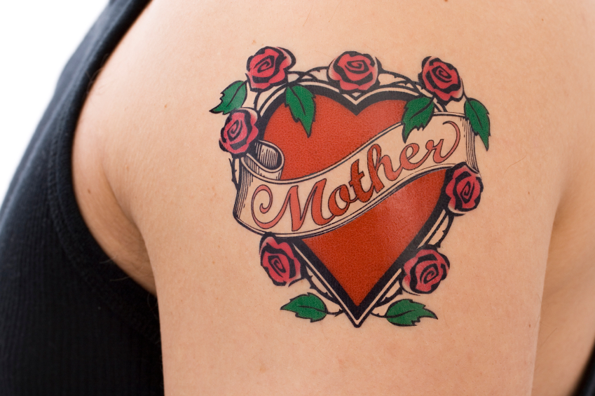 Decorative tattoo on a male arm featuring the word "Mother" on a scroll overlaying a red heart surrounded by pink roses with green leaves.
